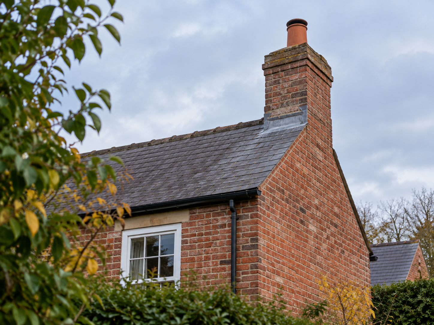 Ground-level view of a traditional red-brick chimney stack with a clay pot, weathered mortar joints and slate roof.
